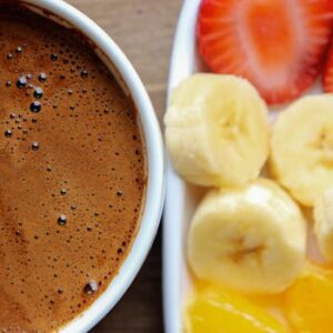 A rich cup of coffee paired with fresh sliced banana, strawberry, and orange on a tray.