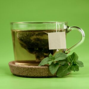 Transparent glass of hot green tea placed on ceramic cup holder with mint leaves on light green surface