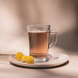 A clear tea glass with brown tea and yellow flowers on a white saucer in soft lighting.
